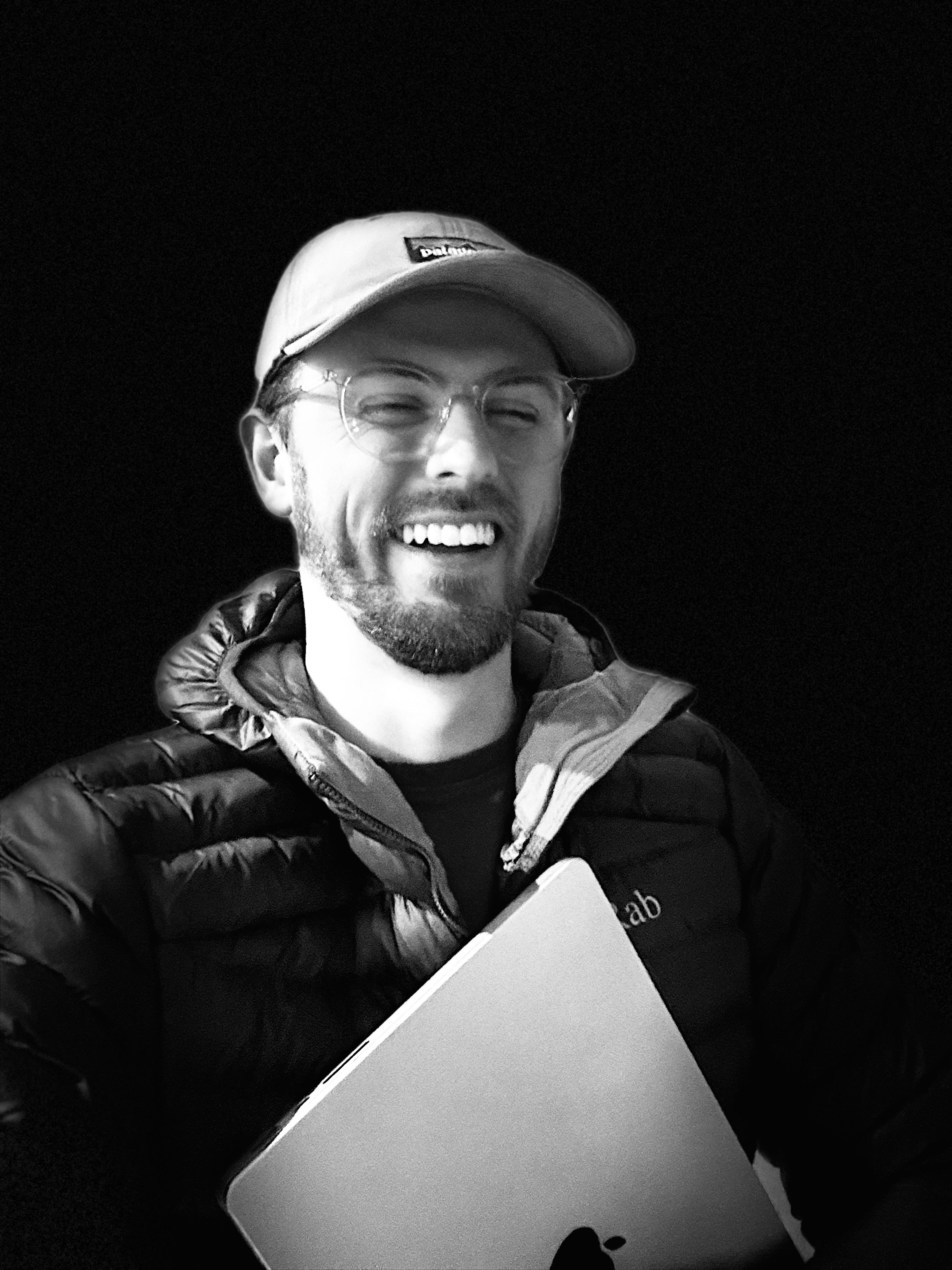 Ben Payne Hunt smiling in a black white wearing a Patagonia hat, Rab coat and clear frame glasses while holding a Macbook